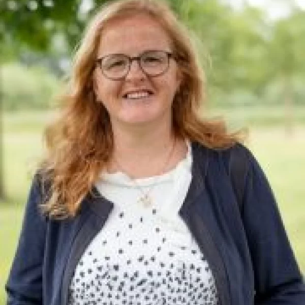Vrouw glimlacht buiten met bomen op achtergrond.